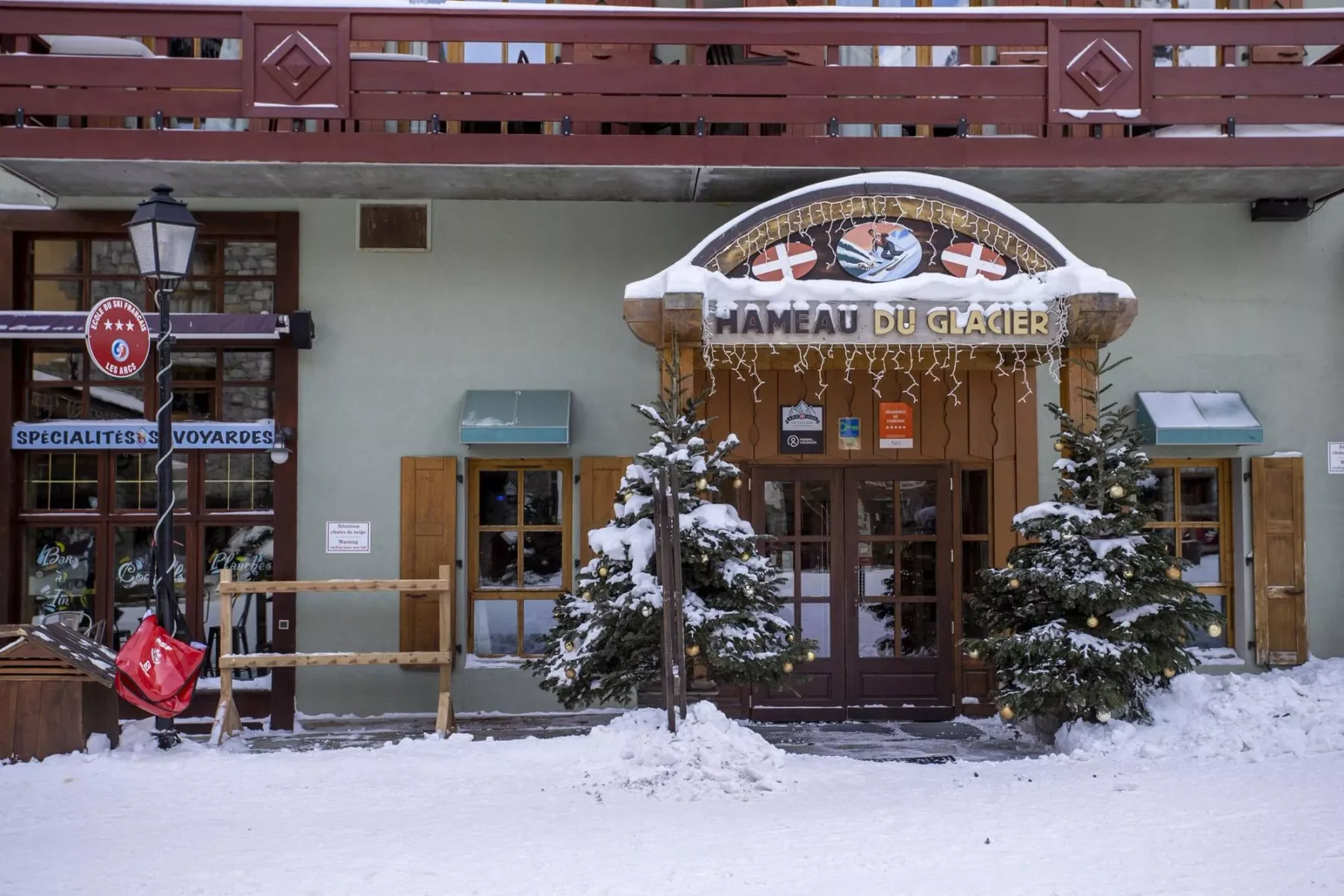 Entrada de un edificio cubierto de nieve, con árboles de hoja perenne y un balcón de madera.
