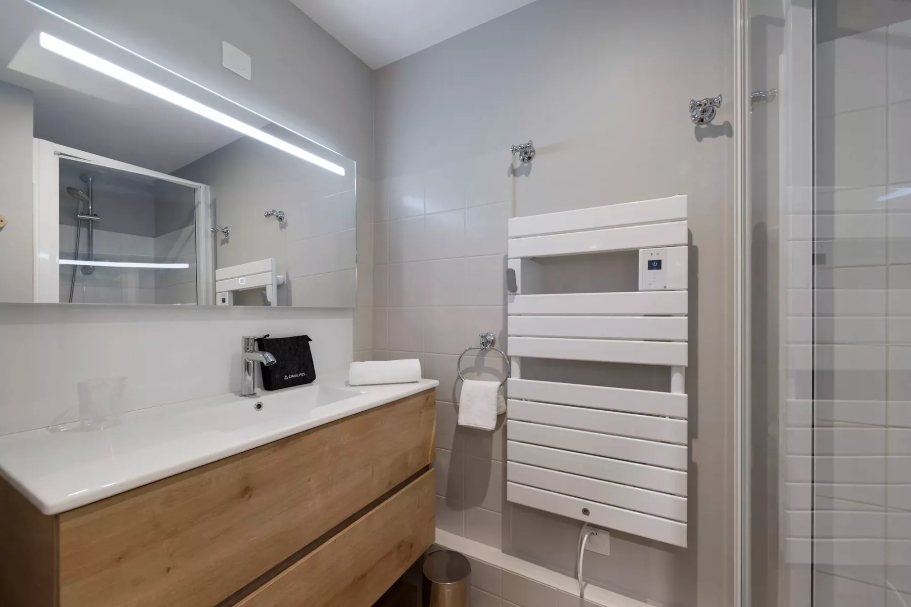 Un baño moderno con un tocador alargado, un radiador toallero mural y una ducha de vidrio.