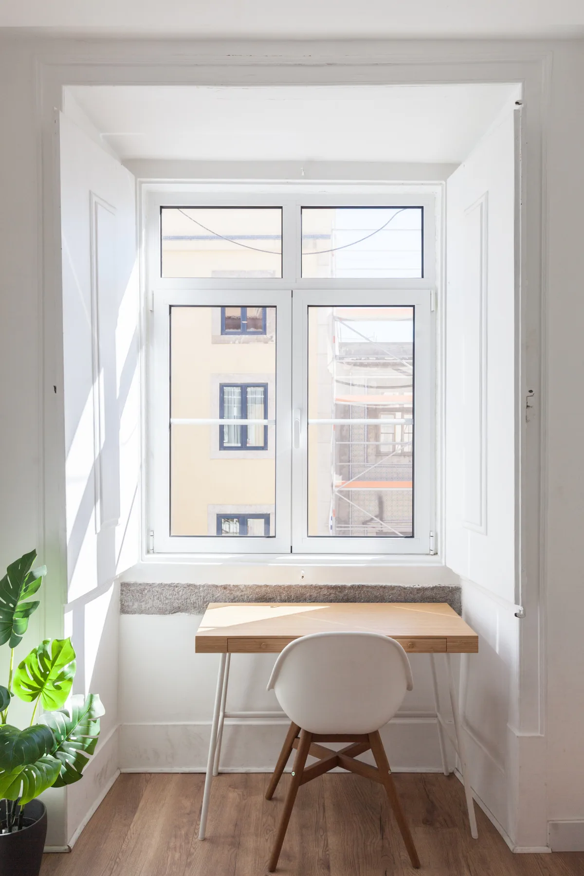 A small desk and chair sit in front of a bright window, with a plant beside them.