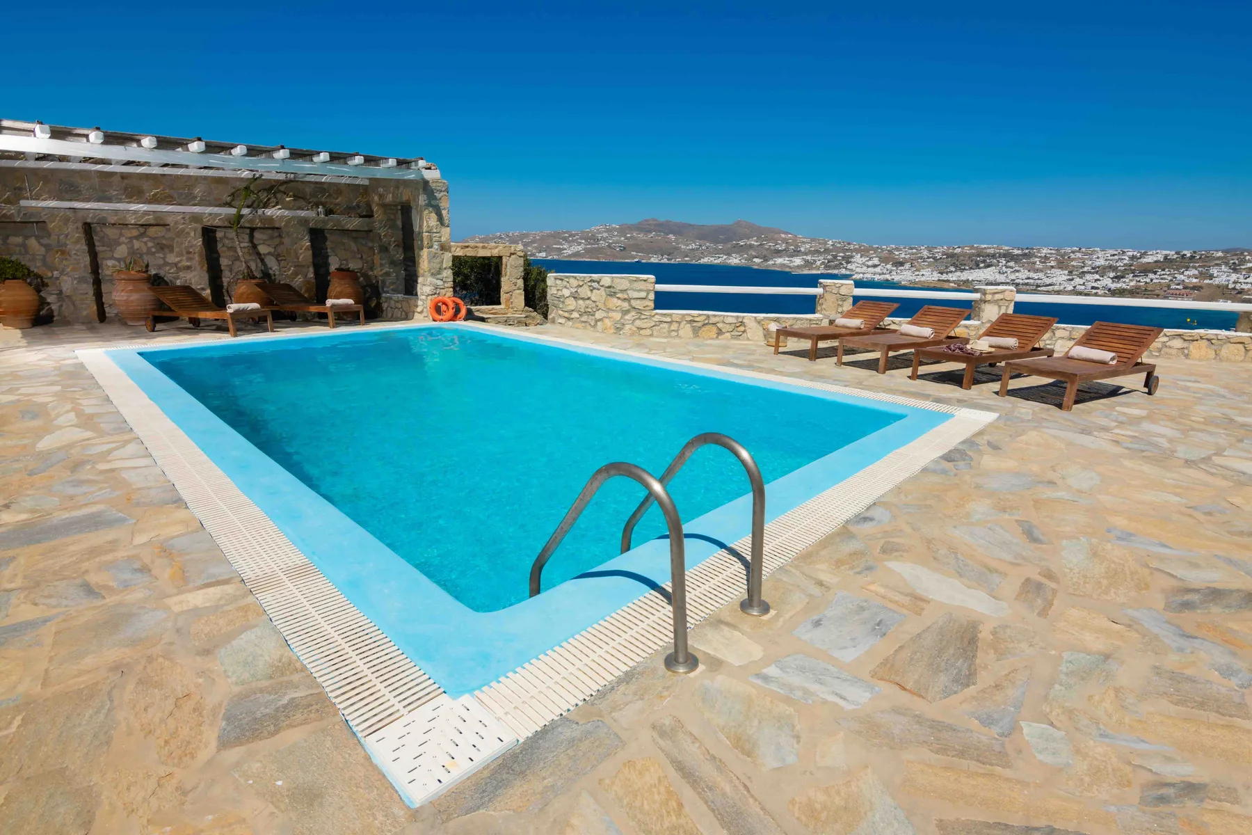 An outdoor swimming pool sits beside a stone terrace with loungers and a sea view.