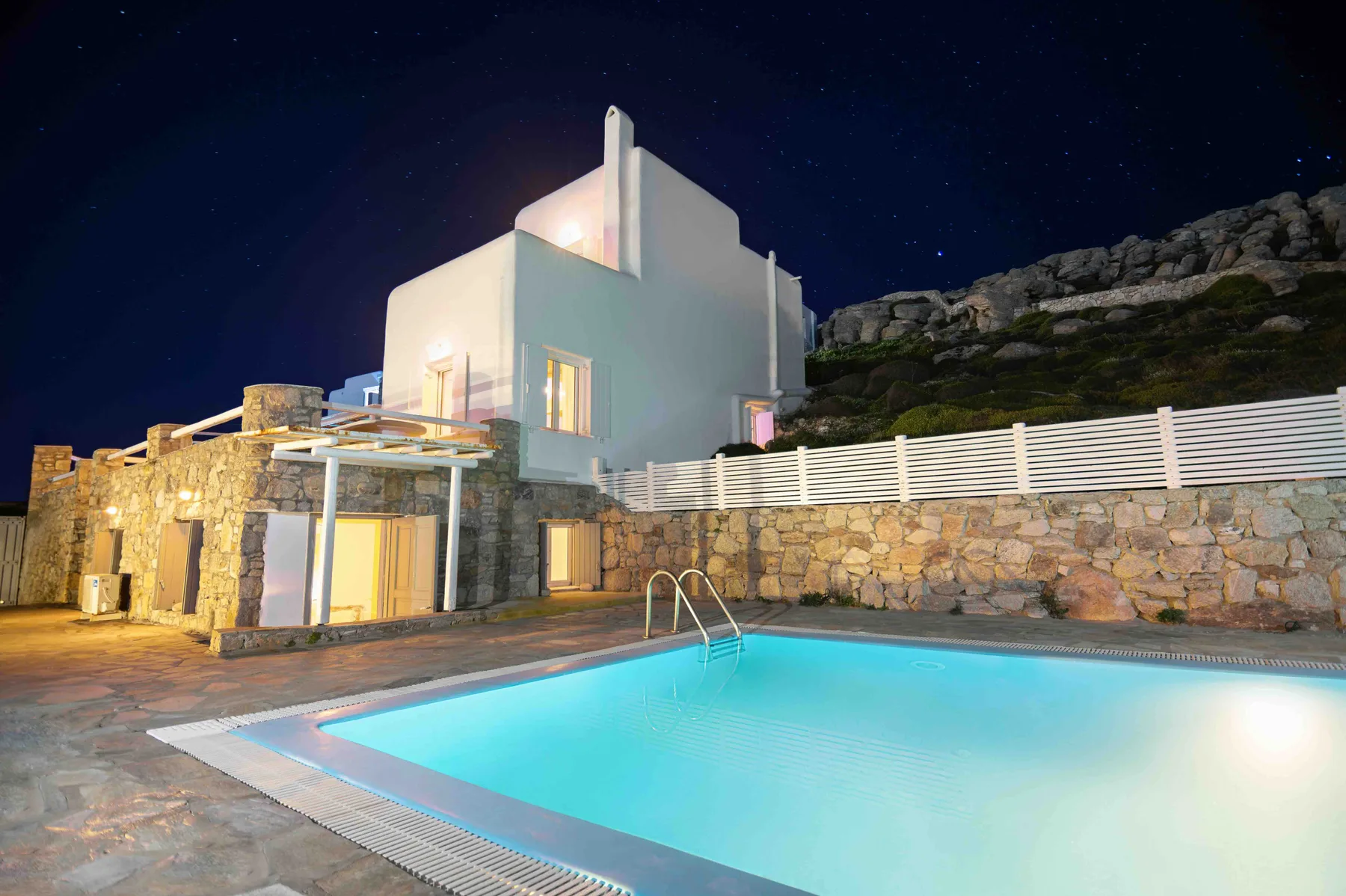 A swimming pool sits beside a white building and stone wall at night.