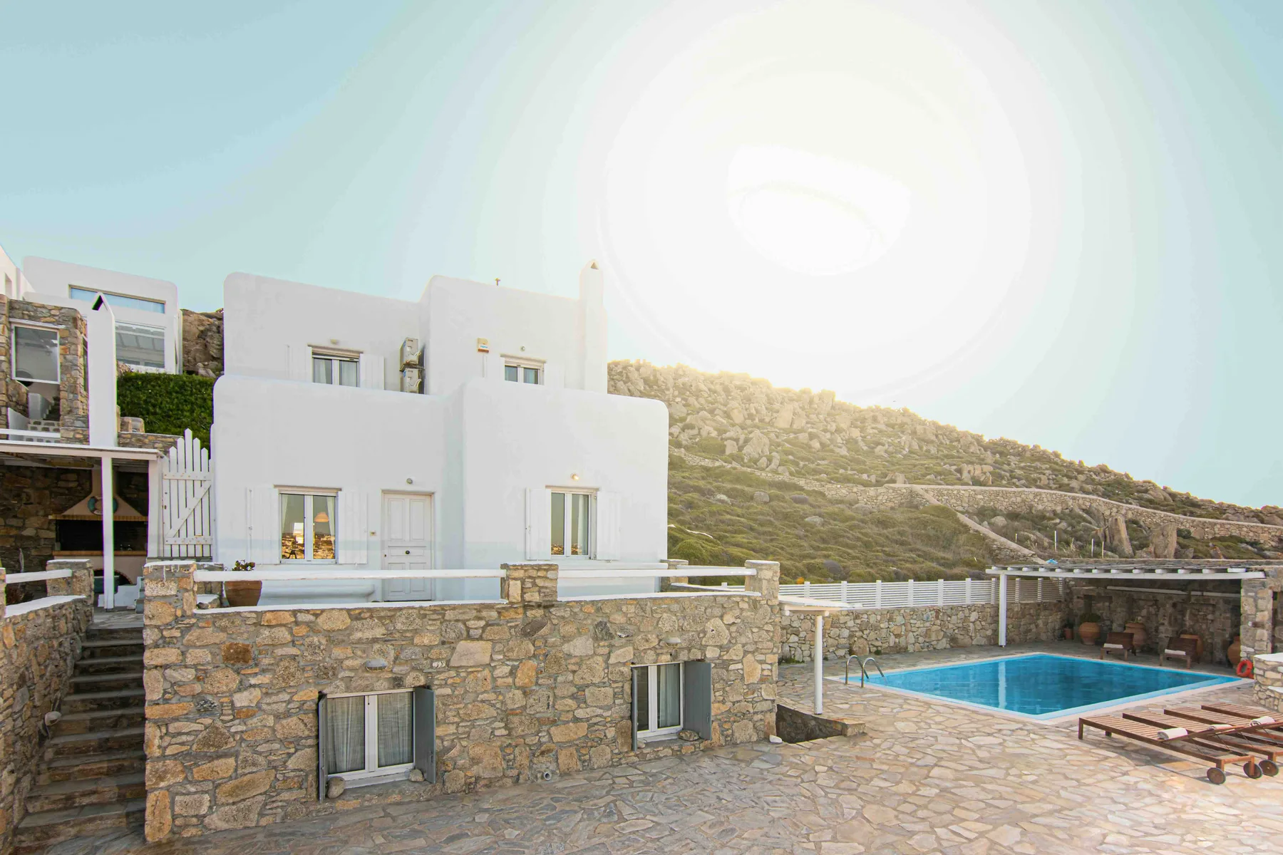 A white villa stands beside a blue swimming pool and stone terrace.