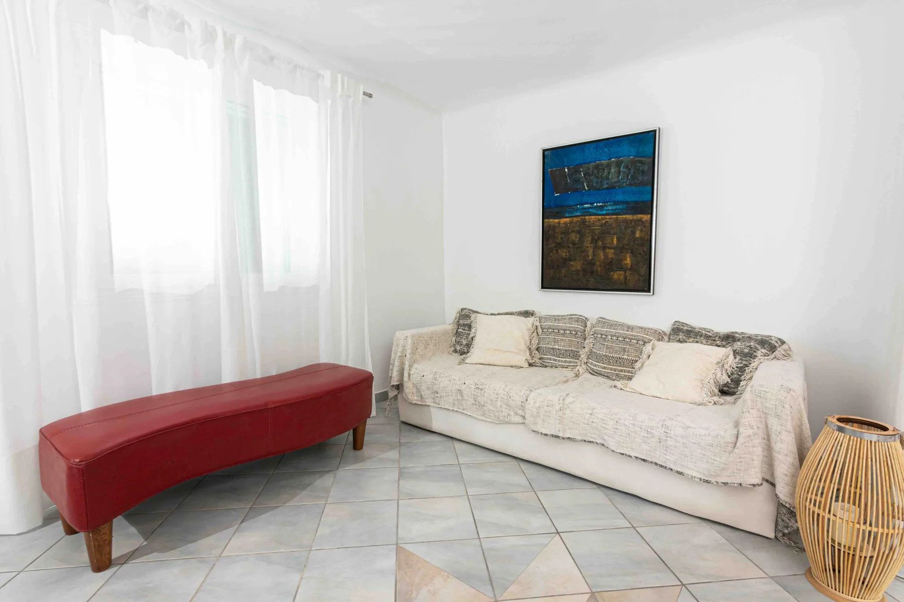 A bright living room with pale seating, a red bench, and sheer curtains.