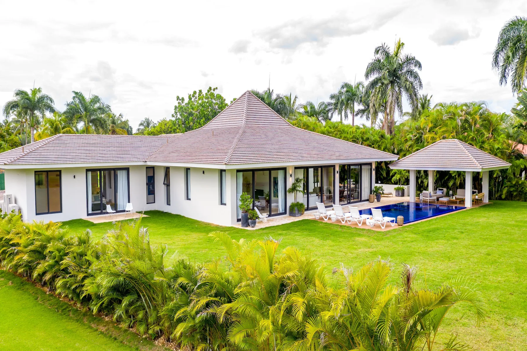 Una casa de una planta y un pabellón cubierto junto a la piscina están rodeados de césped y vegetación tropical.
