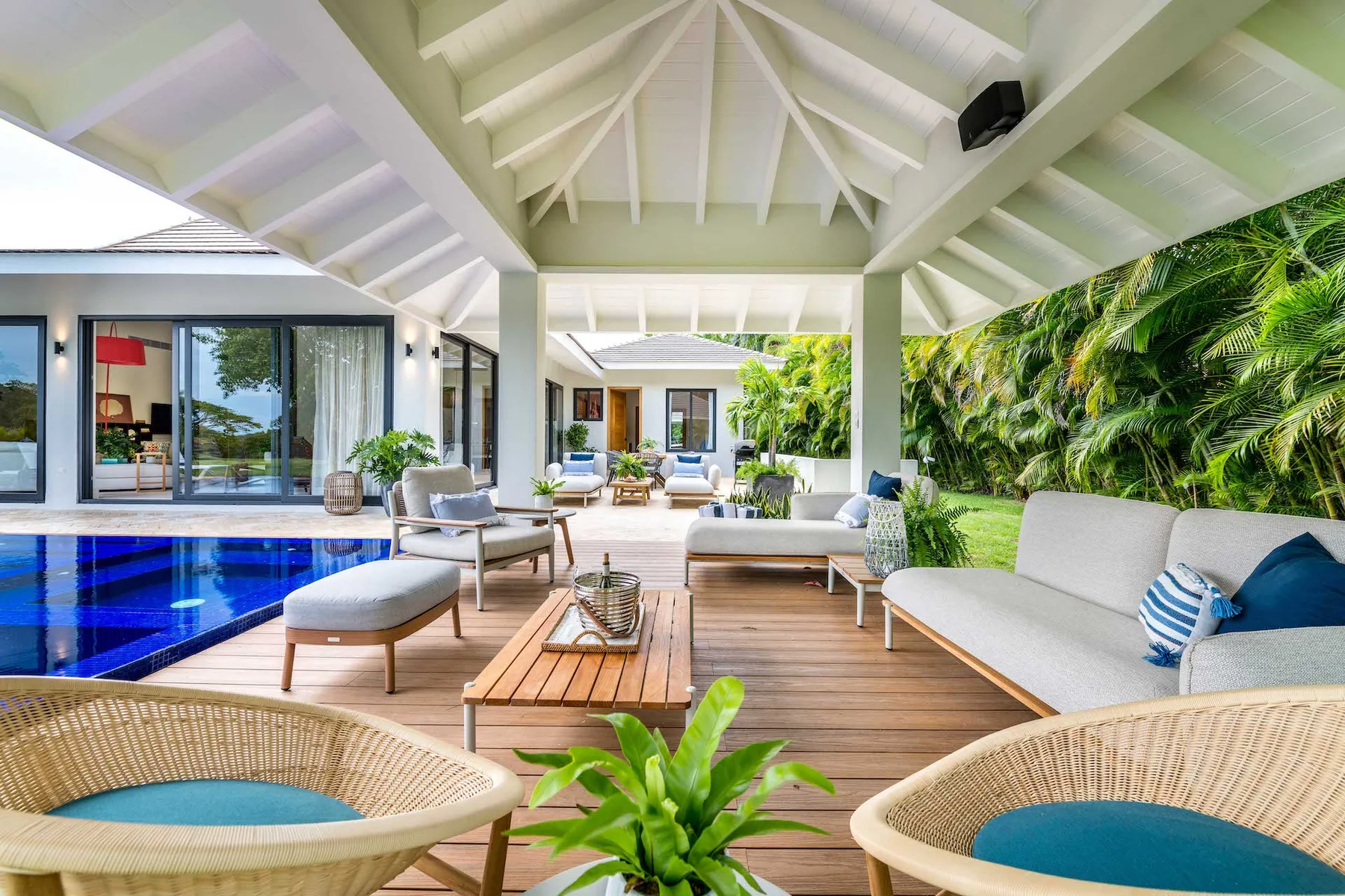 Una sala exterior cubierta se abre a una piscina azul, con asientos bajo un techo blanco.