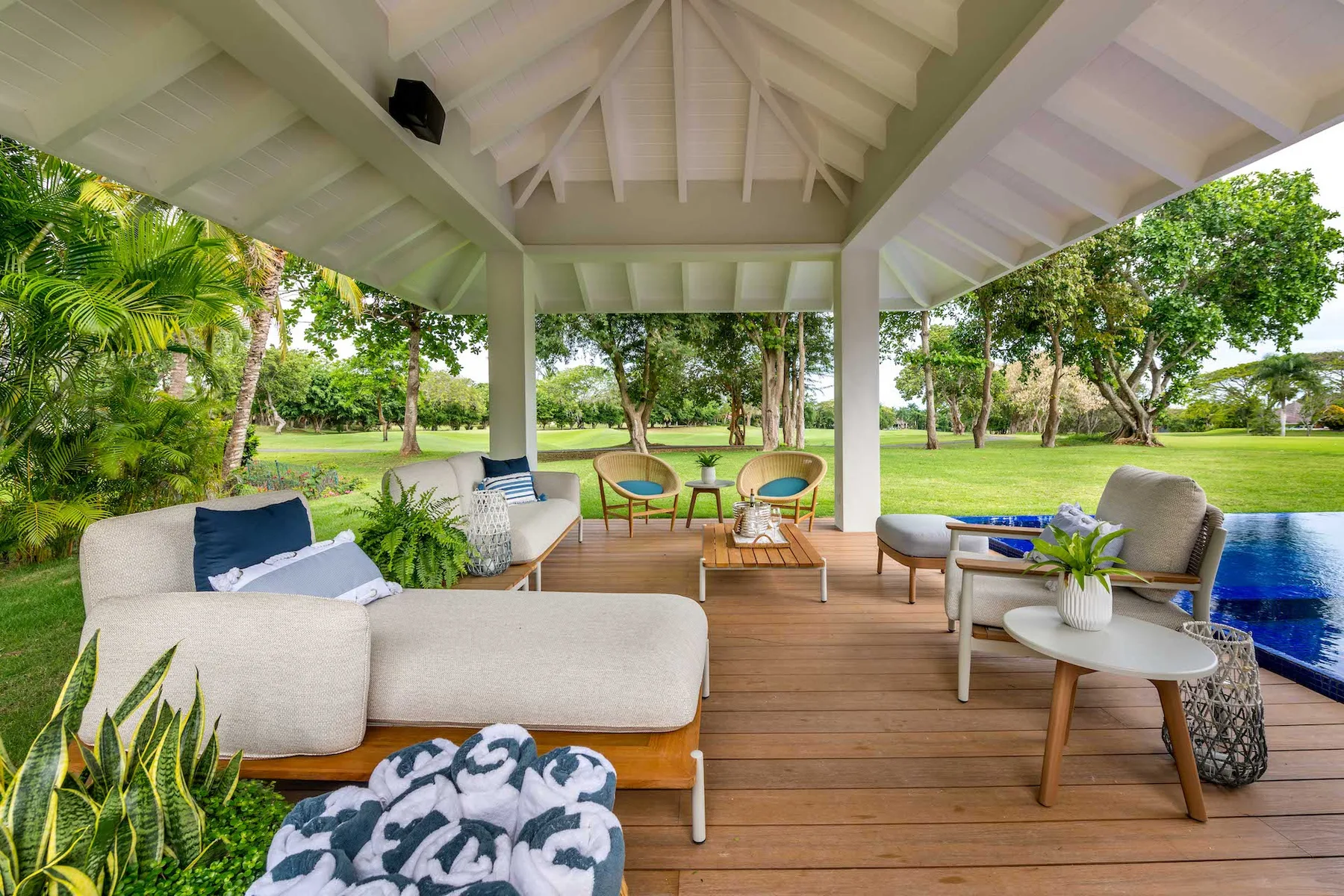 Una sala de estar exterior cubierta con asientos da a un jardín verde y una piscina.