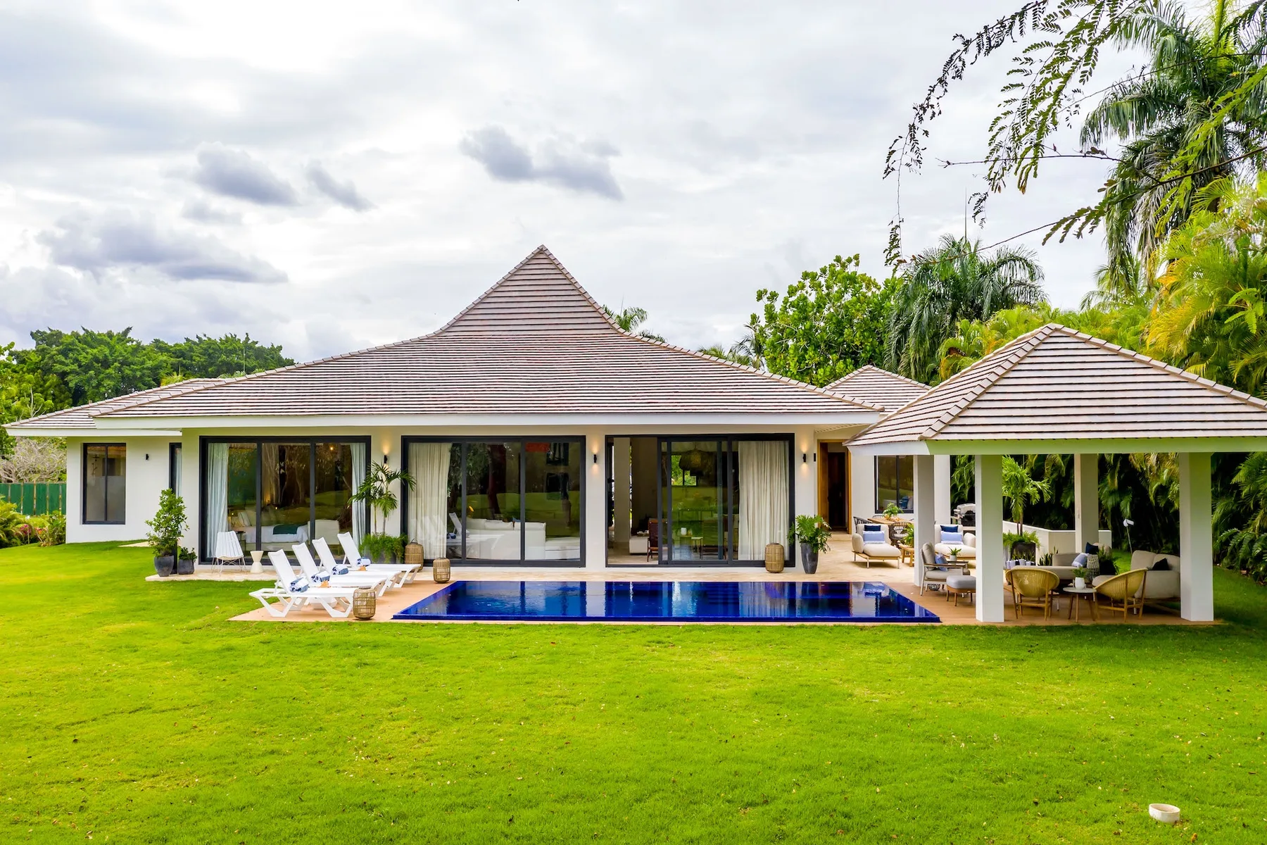 Una casa moderna baja se abre a un césped, con piscina y zona de estar cubierta.
