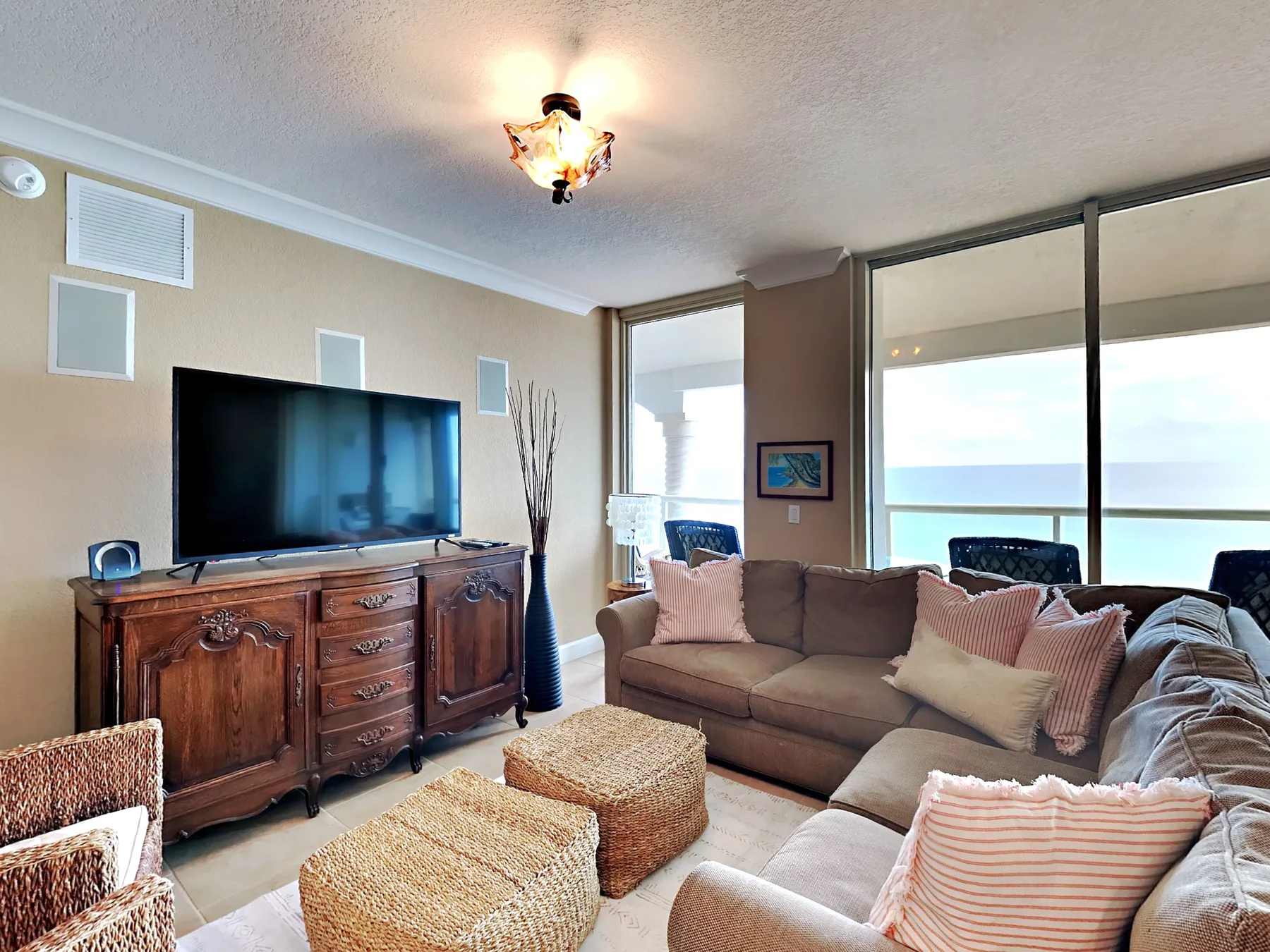 A living room with a television, seating, and woven ottomans beside large windows.
