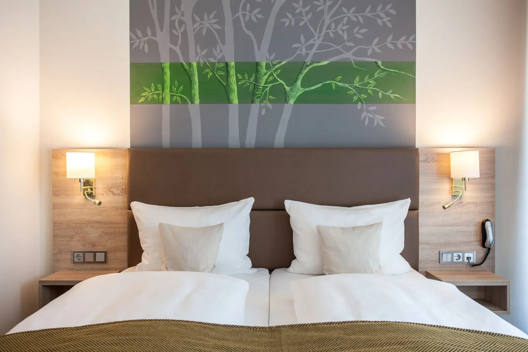 Habitación de hotel con una cama doble, lámparas de noche y decoración mural con motivos de árboles.