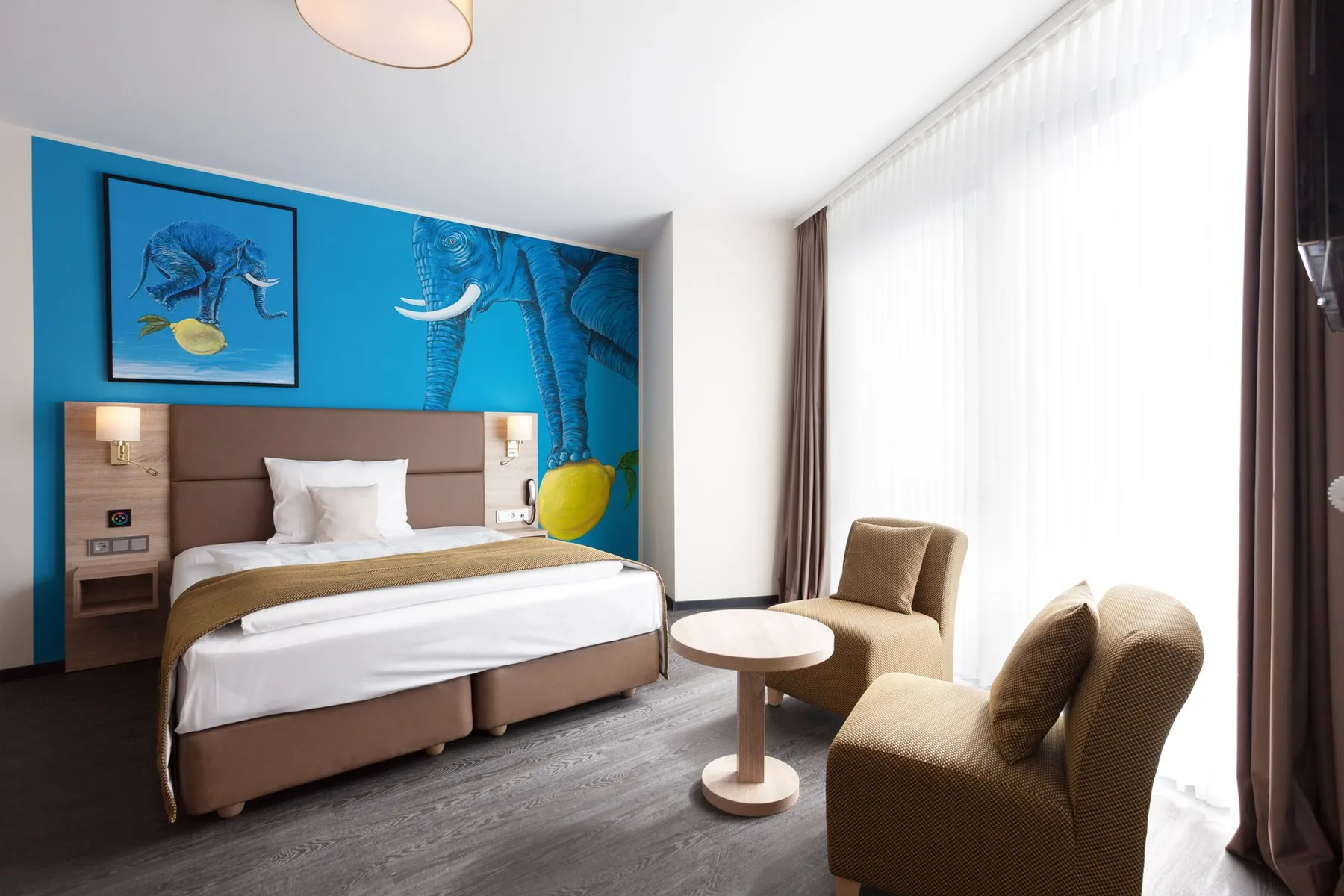 Habitación de hotel con una cama doble, una pared de acento azul, un cuadro de un elefante y dos sillones.