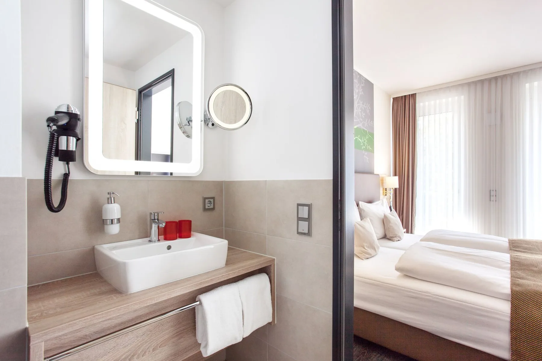 Baño con un lavabo y un espejo, con vistas a un dormitorio con dos camas.