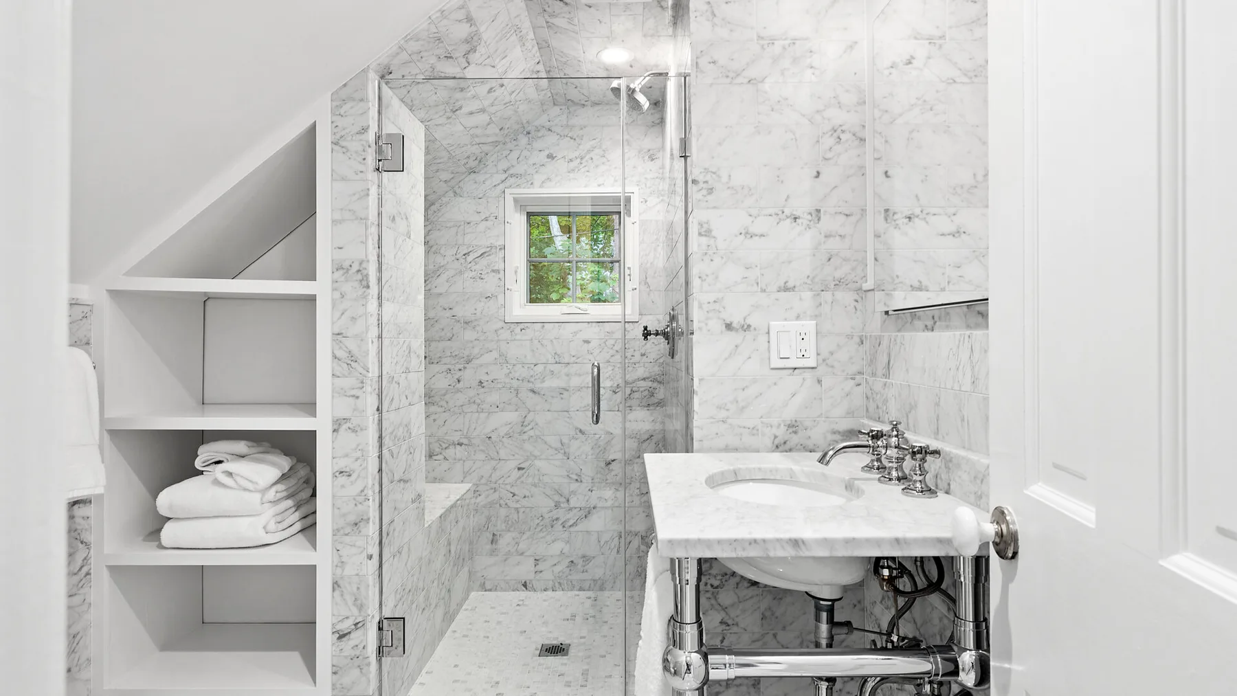 Un baño luminoso de mármol con ducha de vidrio y lavabo con pedestal.
