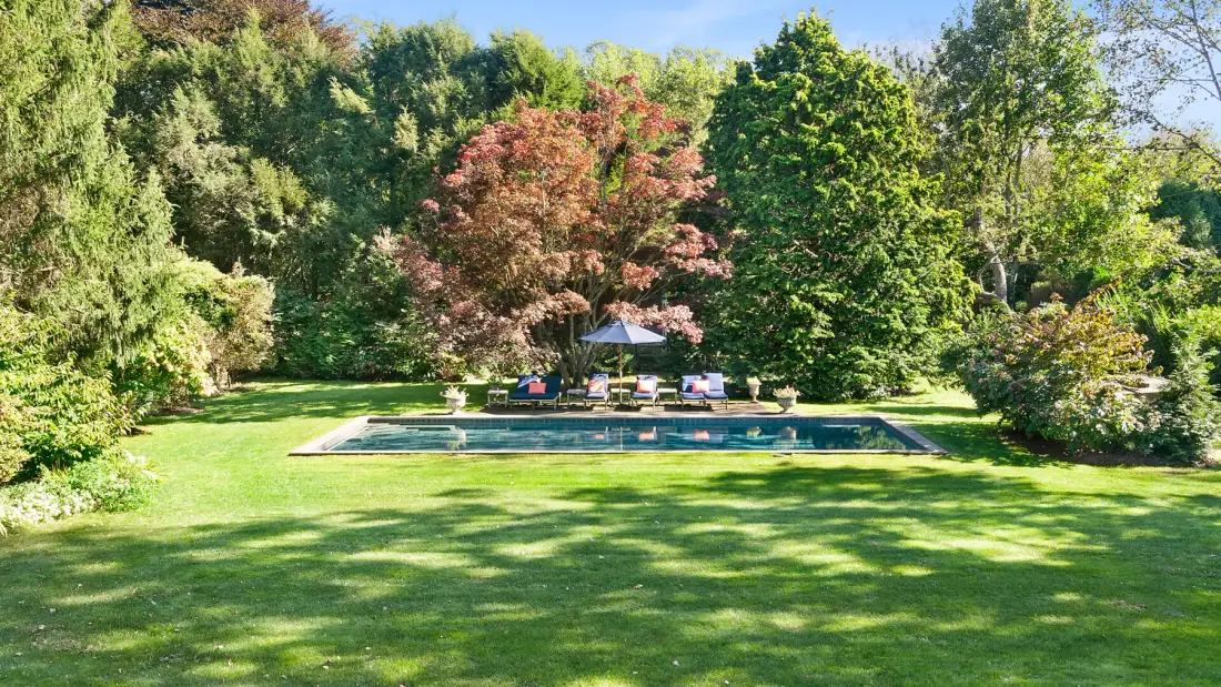 Una piscina se encuentra al fondo de un amplio césped, rodeada de árboles frondosos.