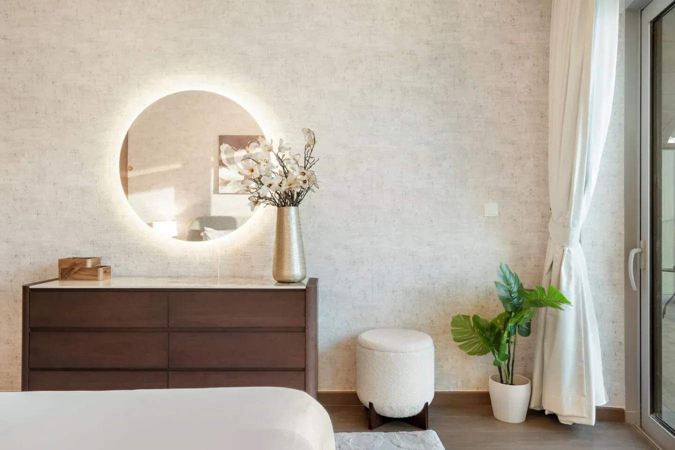 A bedroom with a dark dresser, round mirror, and potted plant beside the window.