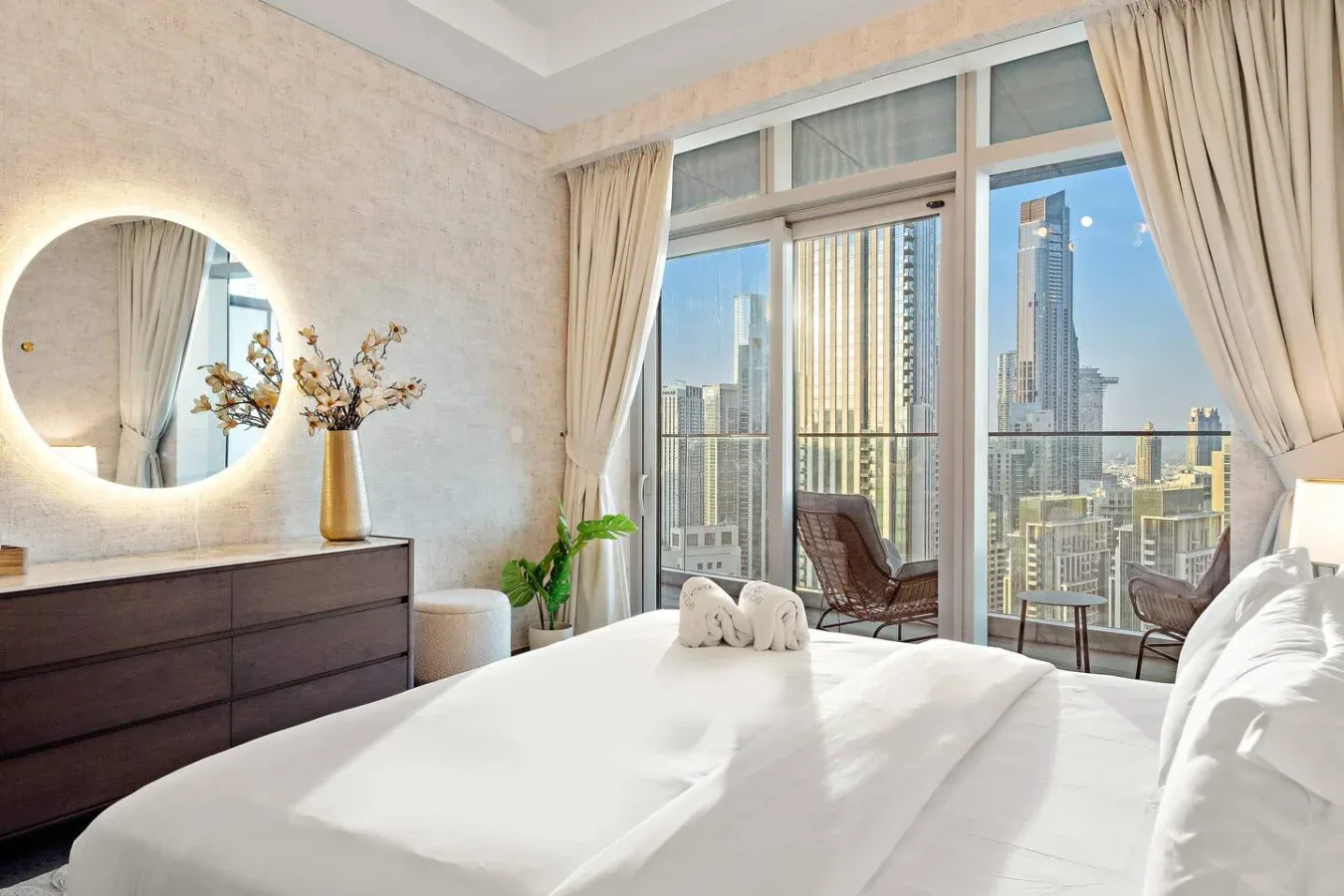 A bright bedroom with a bed beside floor-to-ceiling windows overlooking city buildings.