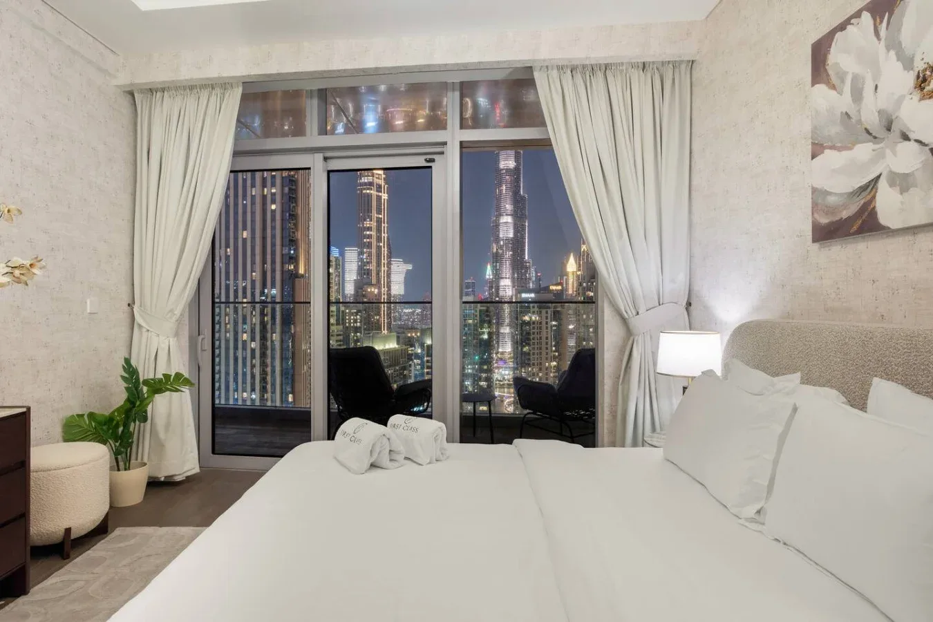 A bedroom with a large bed beside a window overlooking illuminated city buildings.