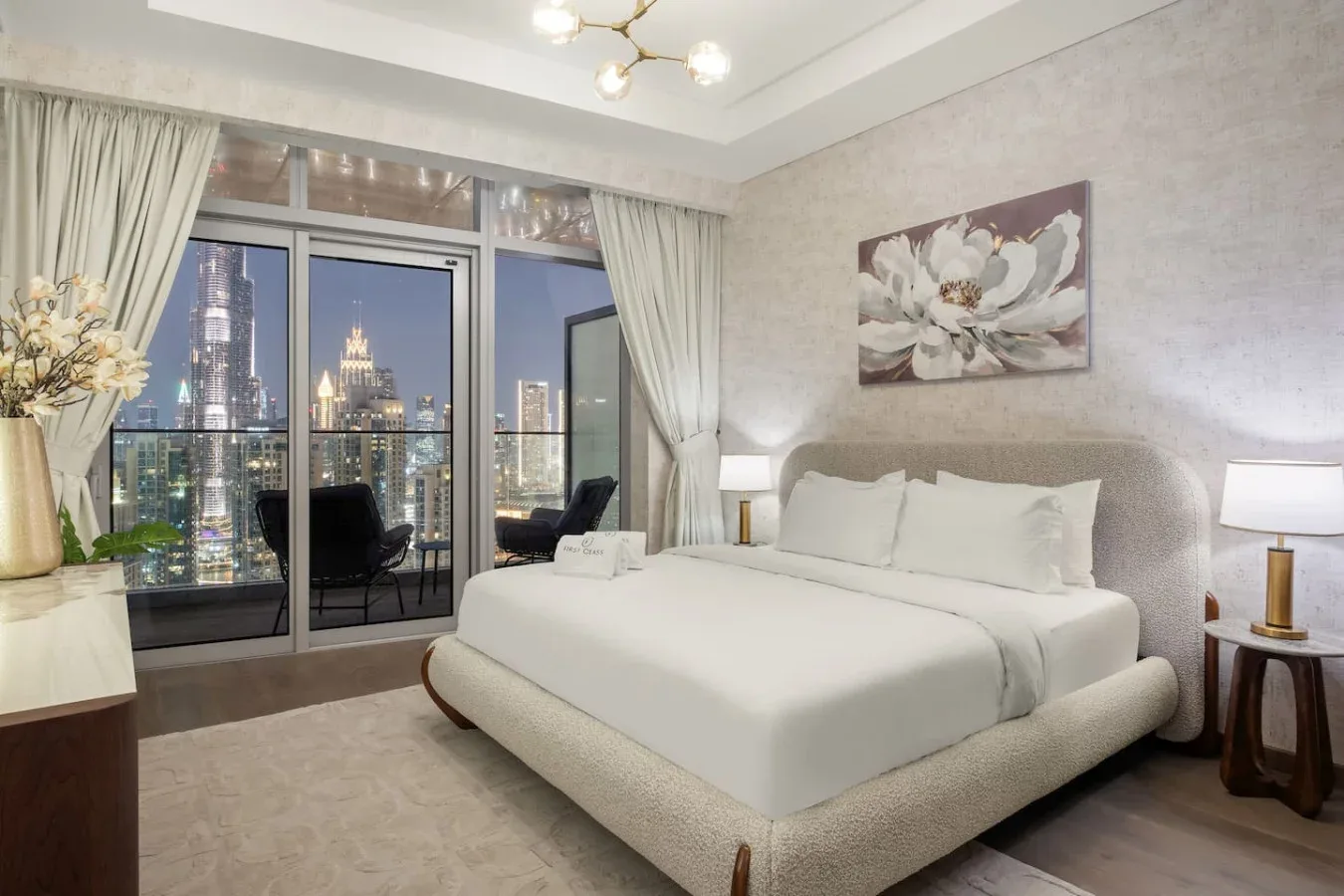 A bright bedroom features a large bed beside floor-to-ceiling windows overlooking city lights.