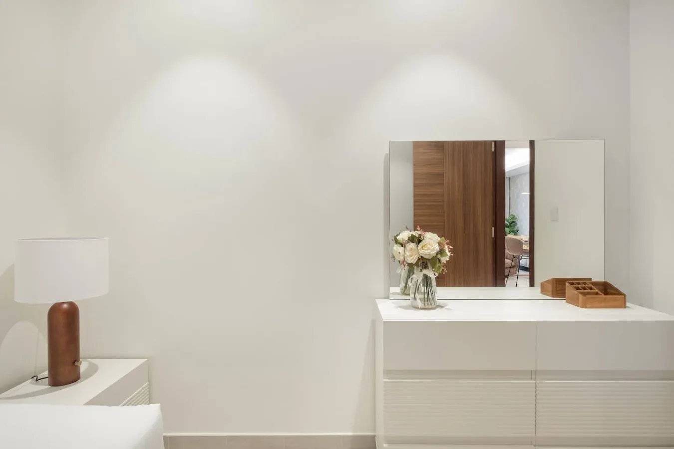 A bright bedroom scene with a bedside lamp and a white dresser topped with flowers.