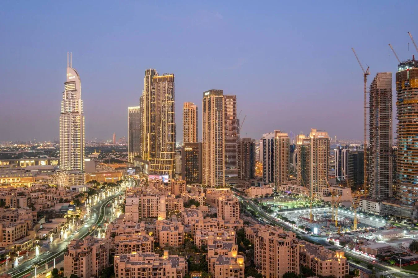 A city skyline rises above dense low-rise buildings and illuminated roads at dusk.