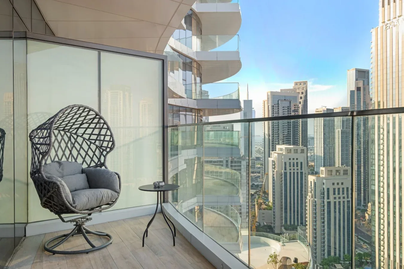 A balcony with a woven lounge chair and glass railing overlooking city high-rises.