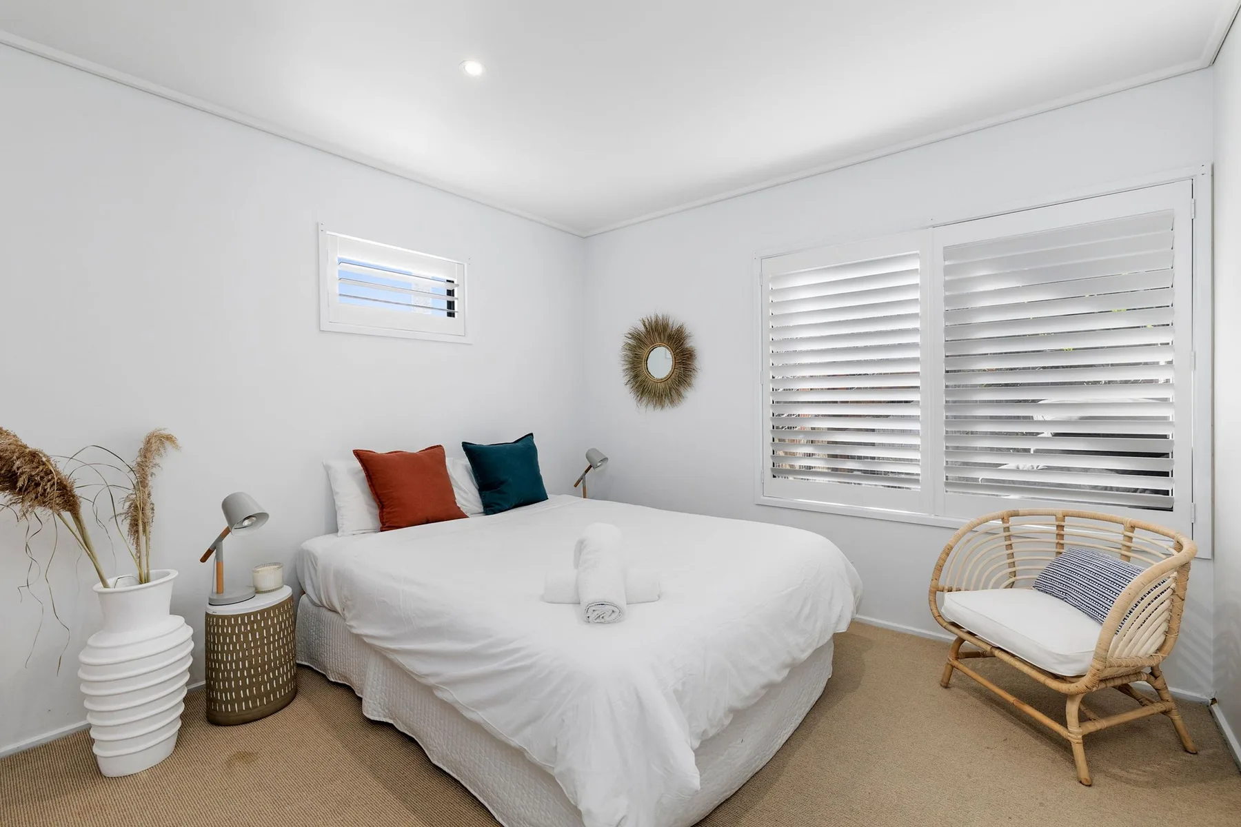 Un dormitorio luminoso con ropa de cama blanca, cojines de colores y una silla tejida.
