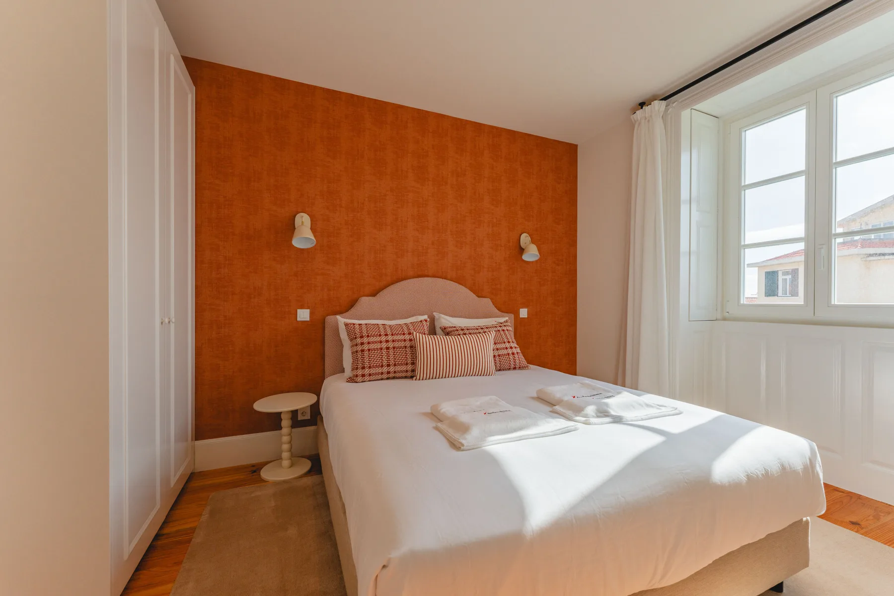 Bedroom with a bed against an orange accent wall beside a large window.