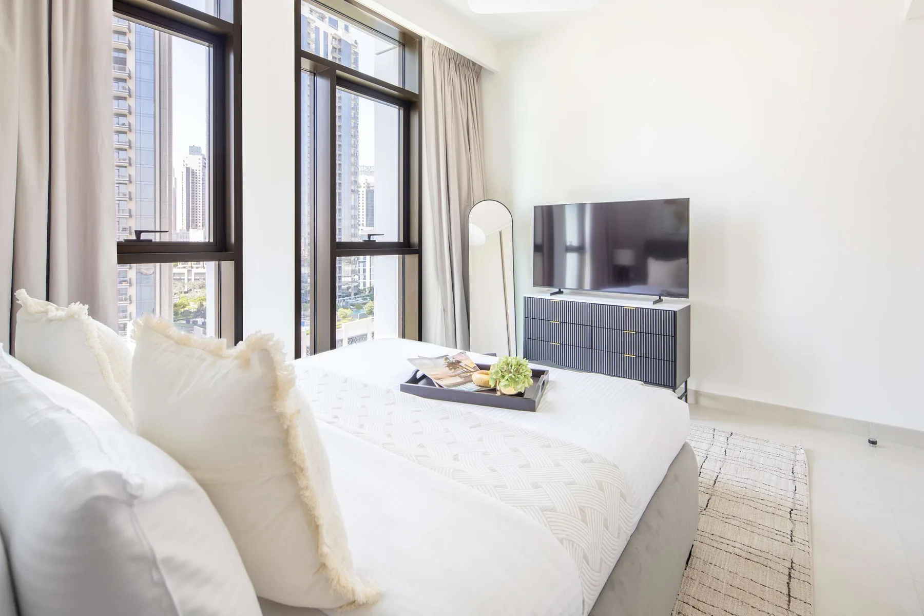A bright bedroom with a bed beside tall windows and a television.