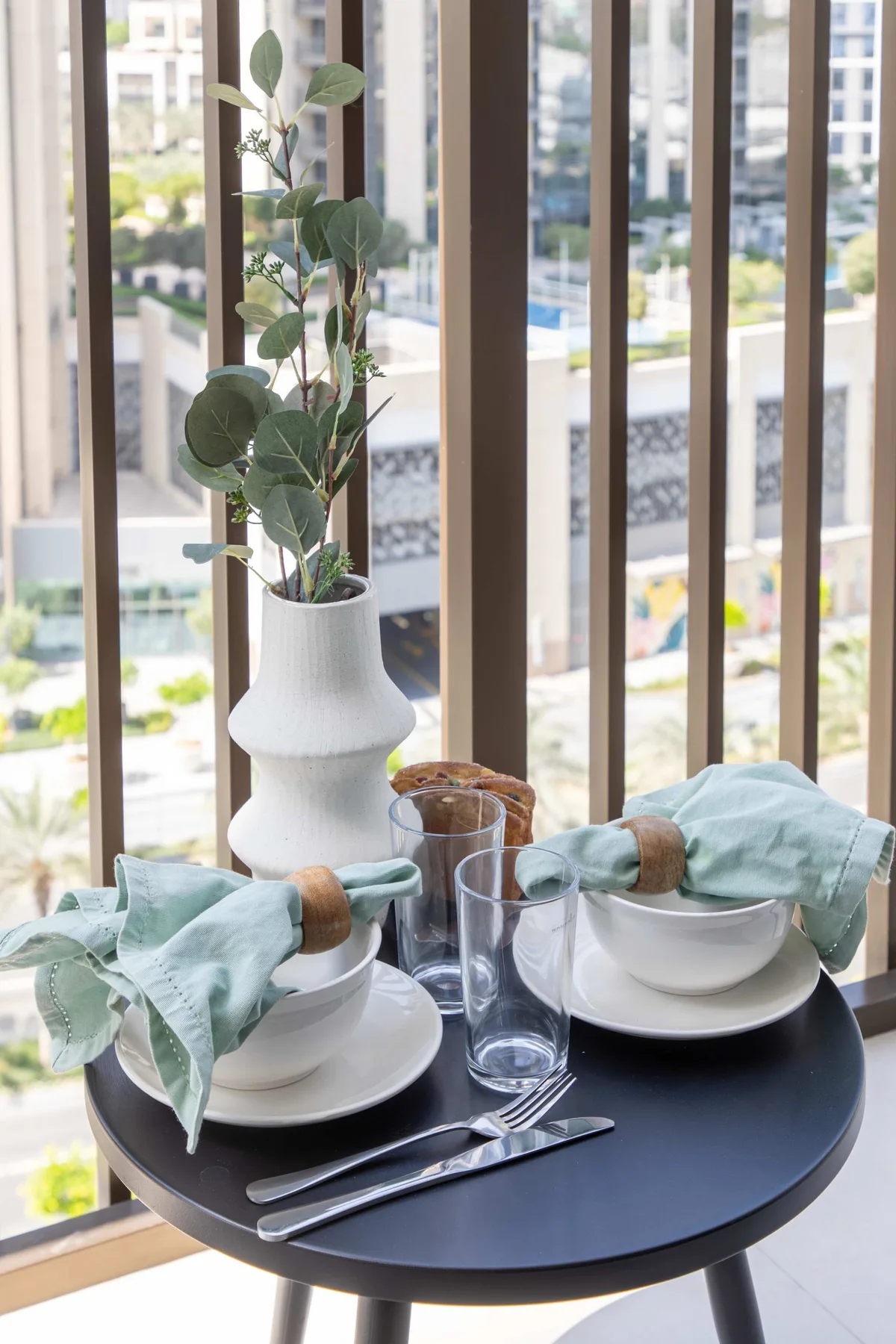 A small dining table is set with plates and glasses beside tall windows.