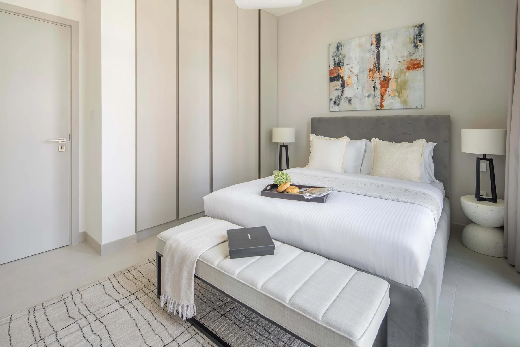 A bedroom with a neatly made bed, a cushioned bench, and abstract wall art.