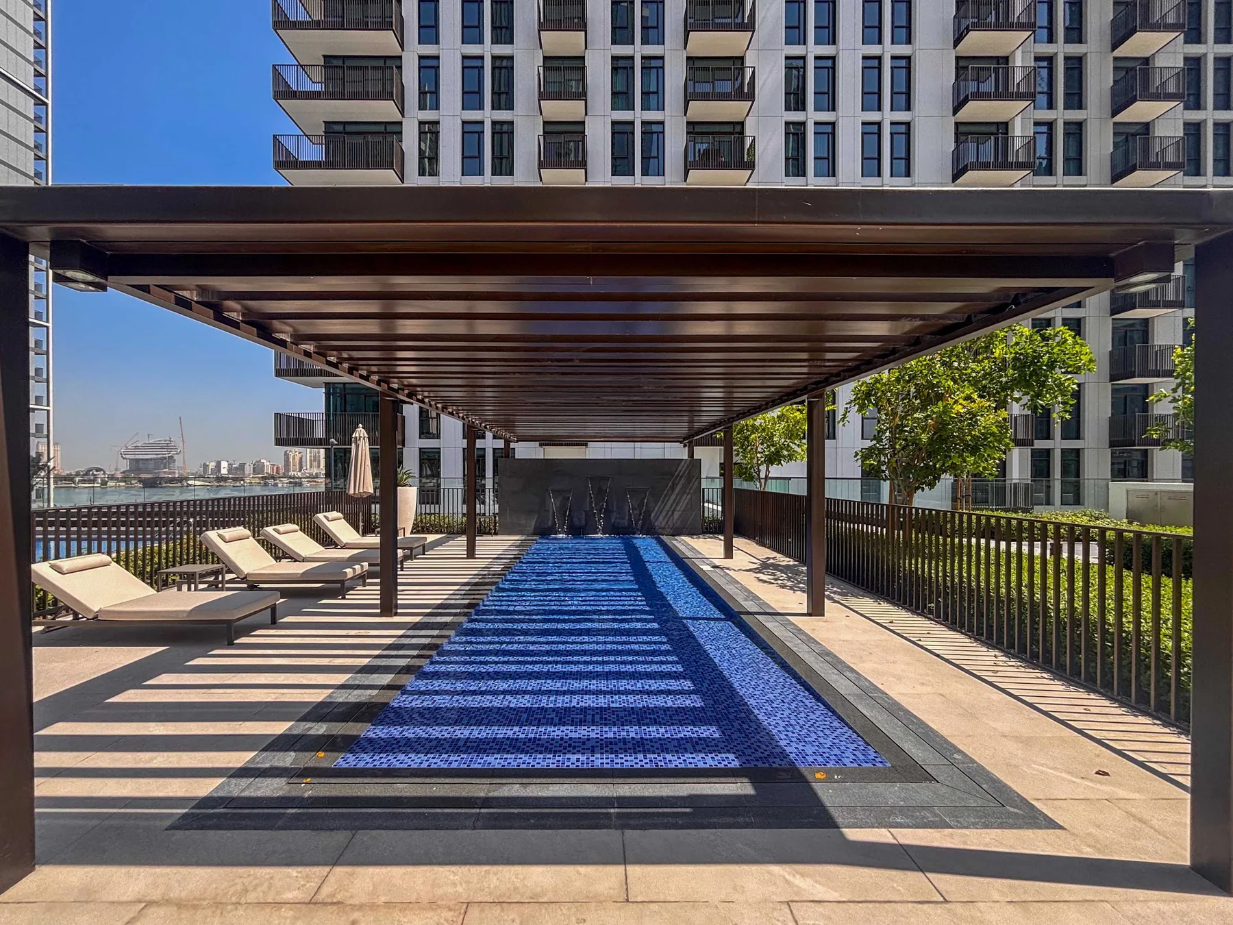 A long outdoor swimming pool sits beneath a shaded pergola beside an apartment building.