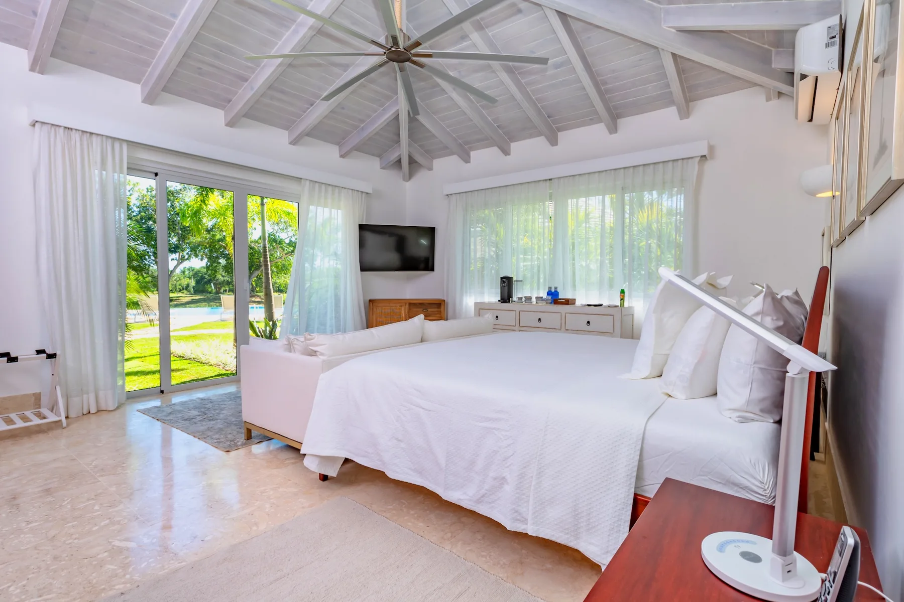 A bright bedroom with white bedding, a vaulted ceiling, and garden-facing windows.