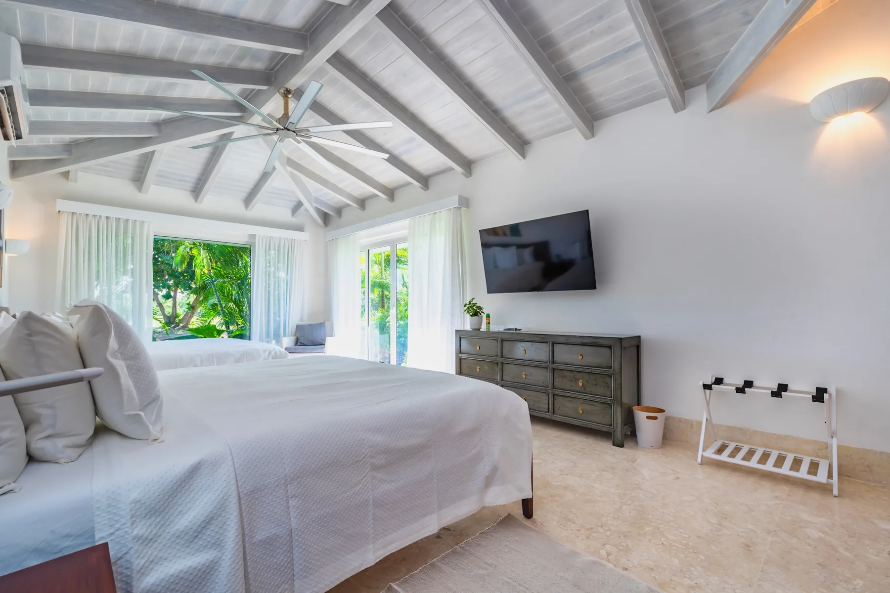 A bright bedroom with a large bed, a wall-mounted television, and windows overlooking greenery.