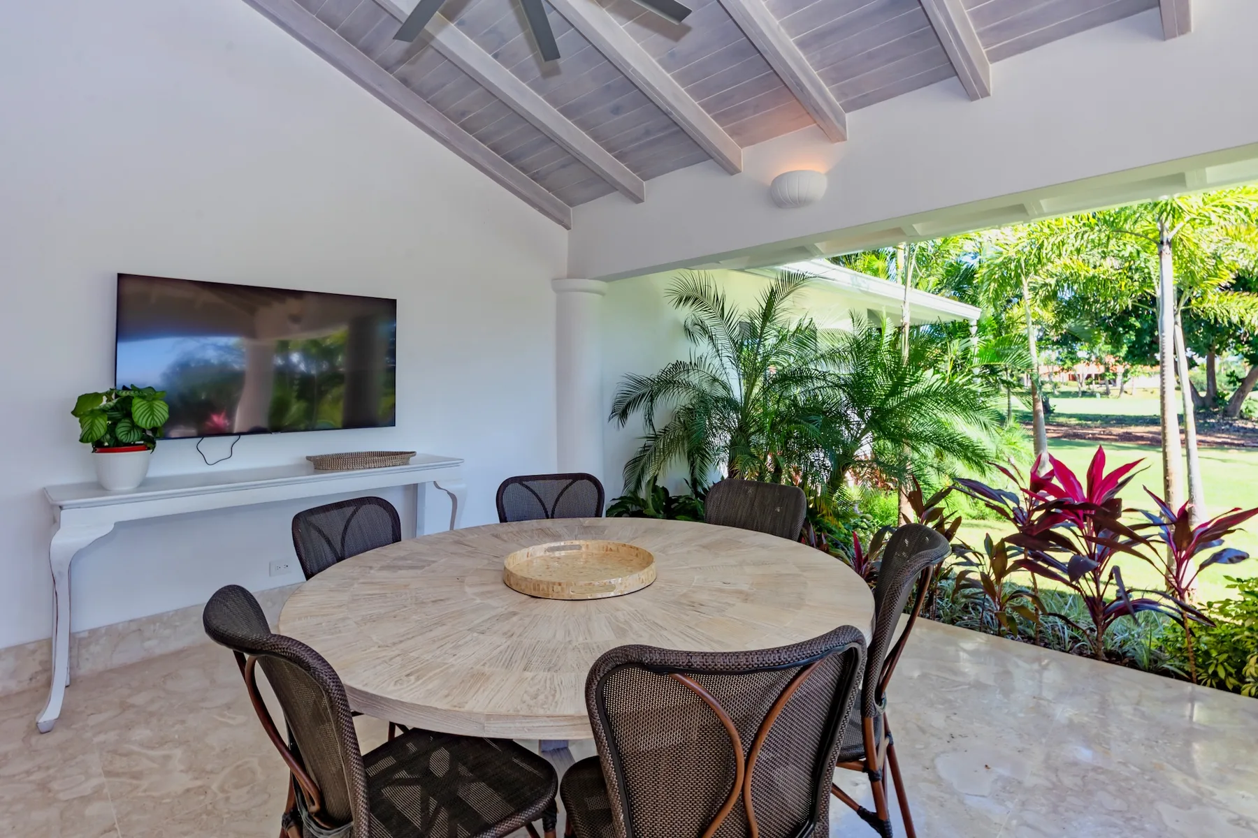 A covered dining area with a round table and chairs, open to a lush garden.