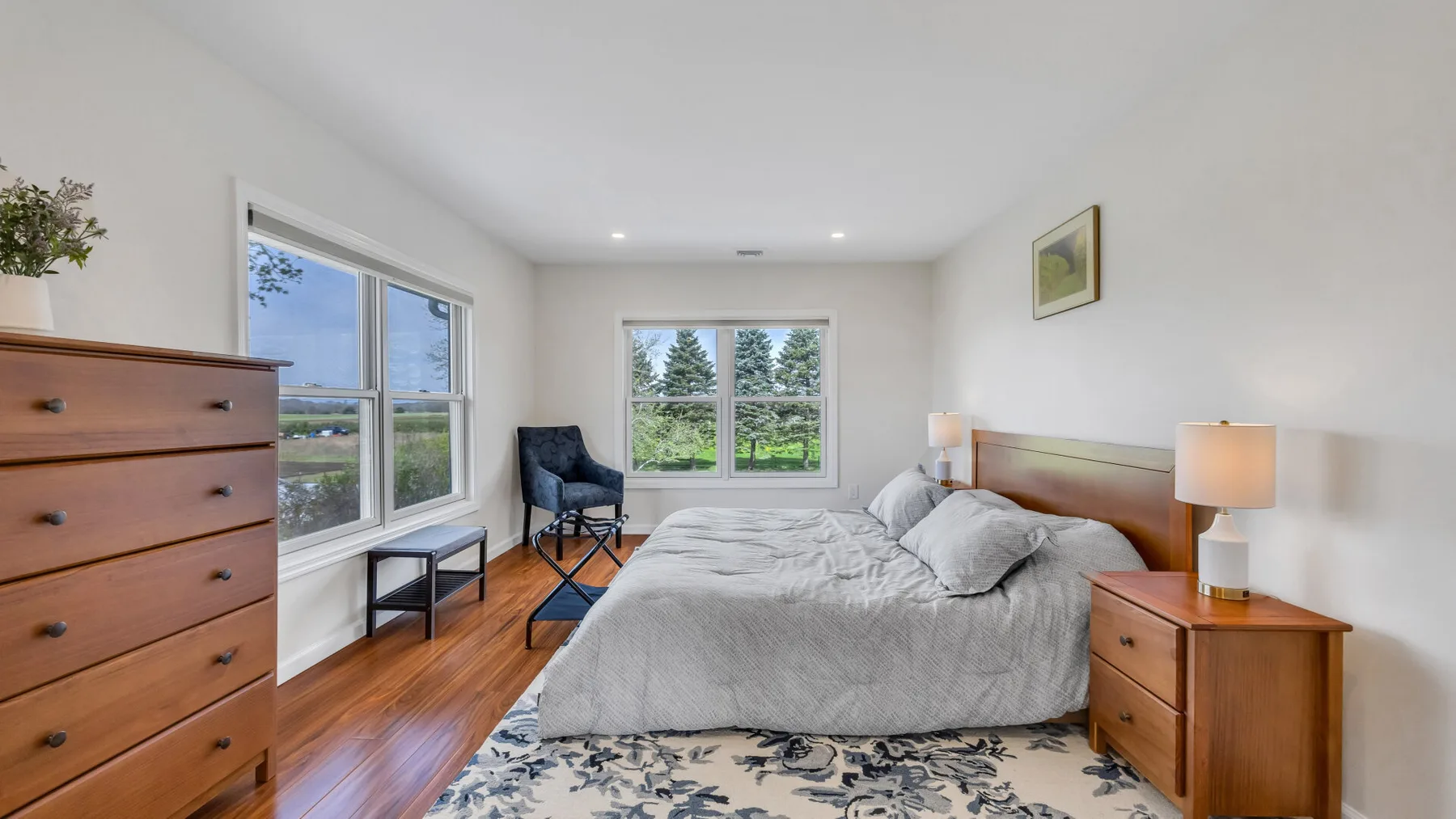 A bright bedroom with a bed, bedside tables, and windows overlooking greenery.