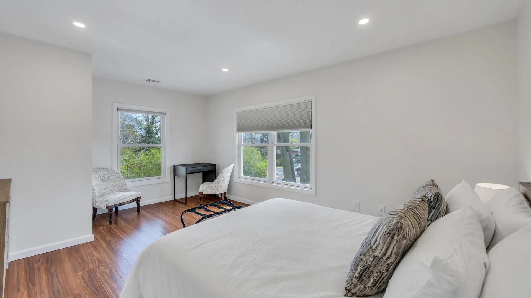 A bright bedroom with a bed, two windows, and wood flooring.