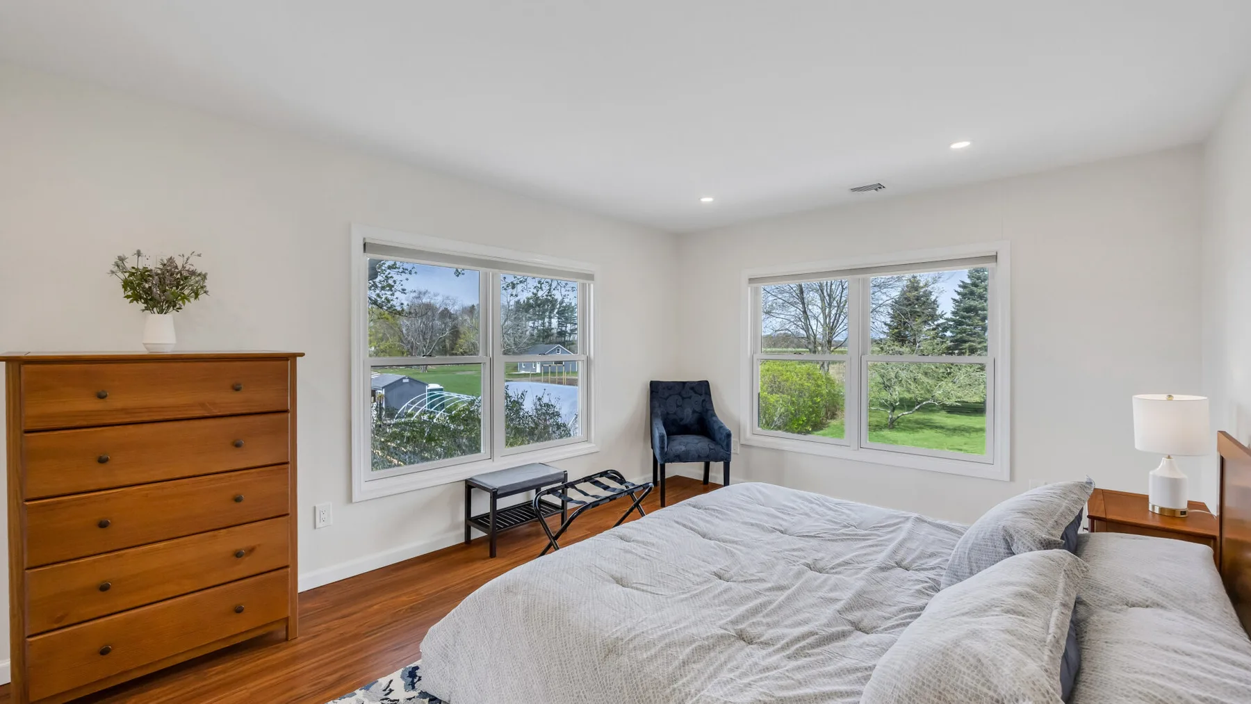 A bright bedroom with a bed, wooden dresser, and large windows overlooking greenery.