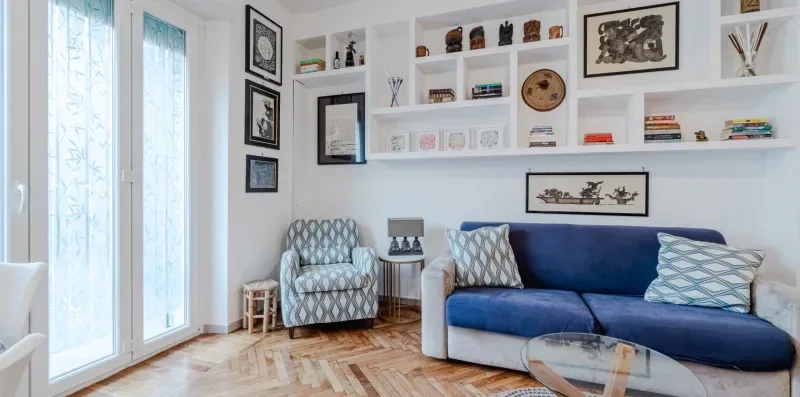 A bright living room with a blue sofa, patterned armchair, and wall shelves.