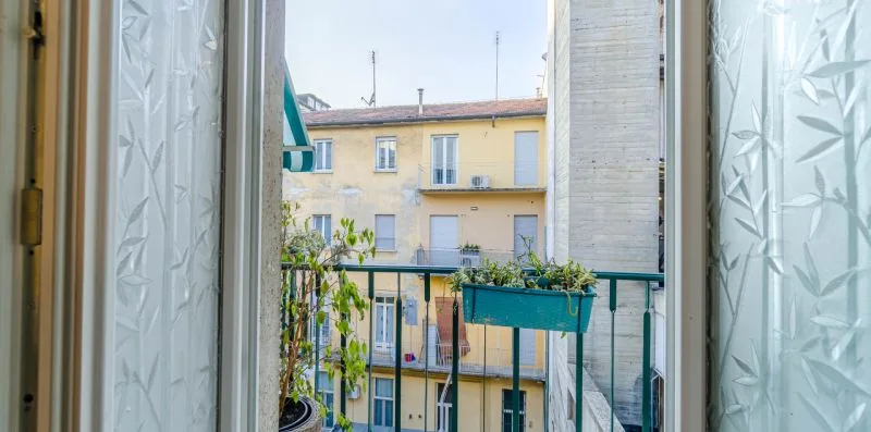An open window looks onto a small balcony with potted plants and a neighboring building.