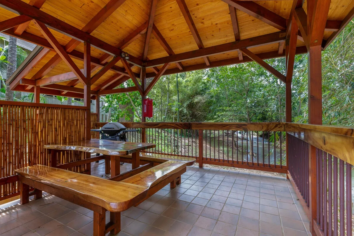 Un comedor exterior cubierto de madera con mesas de picnic y árboles verdes al fondo.