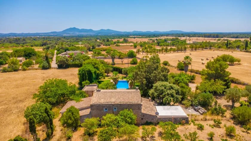 An aerial view shows a house amid fields and scattered trees, with mountains in the distance.