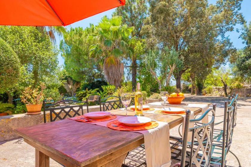 An outdoor dining table is set beneath an orange umbrella in a leafy garden.