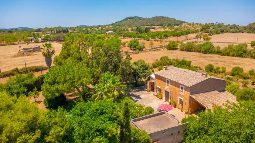 A rural house sits among trees beside open fields and low hills.