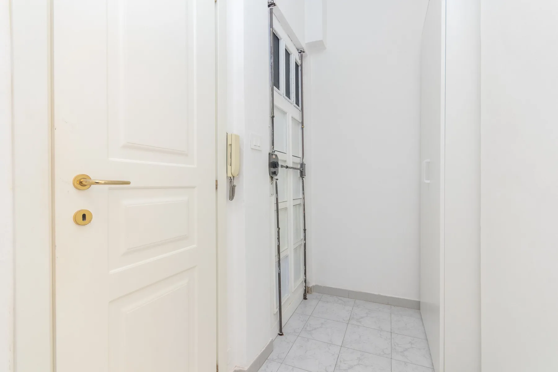 Un pasillo blanco y luminoso con puertas con paneles y suelo de baldosas gris claro.