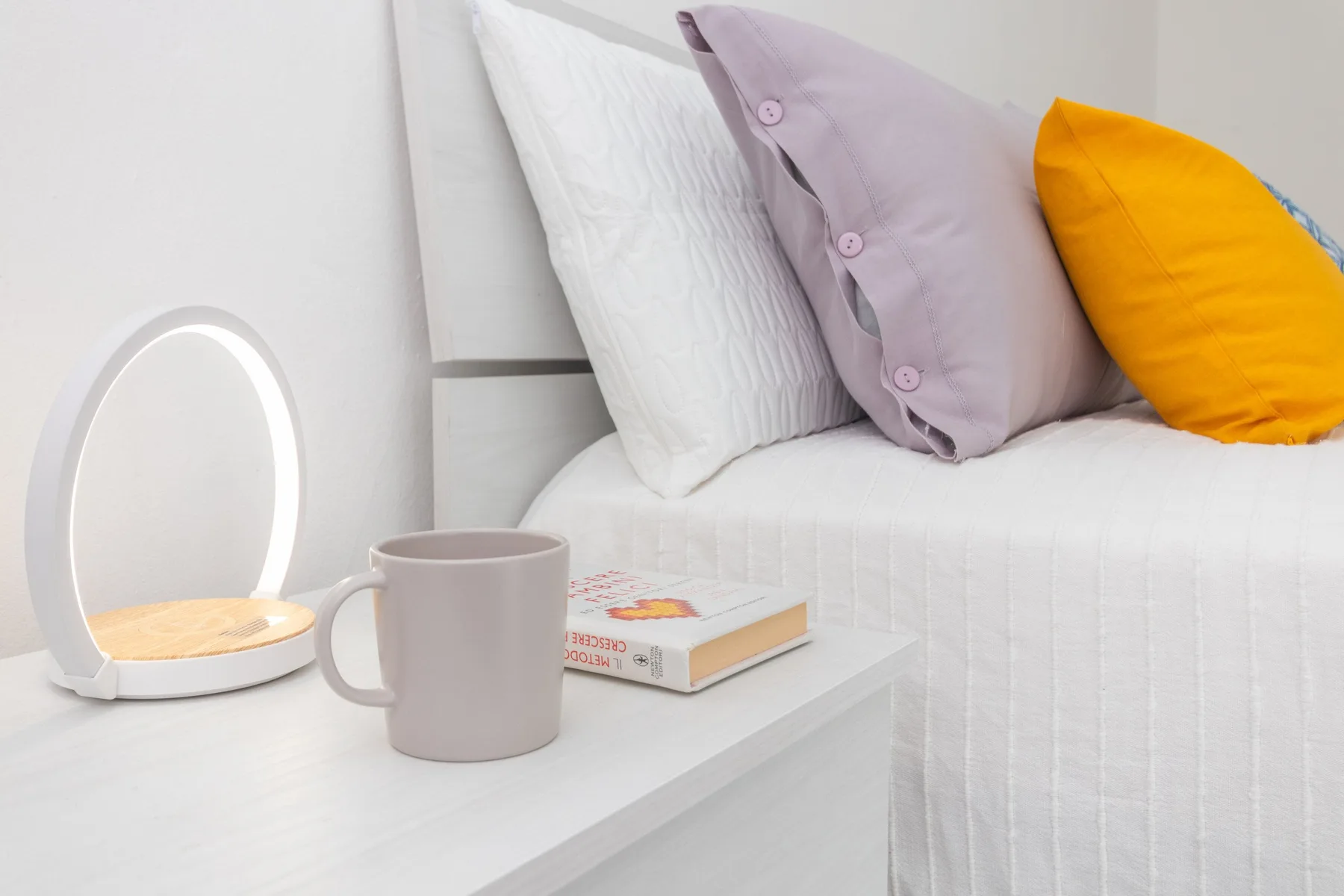 Una cama con almohadas de colores está junto a una mesita con una taza y un libro.