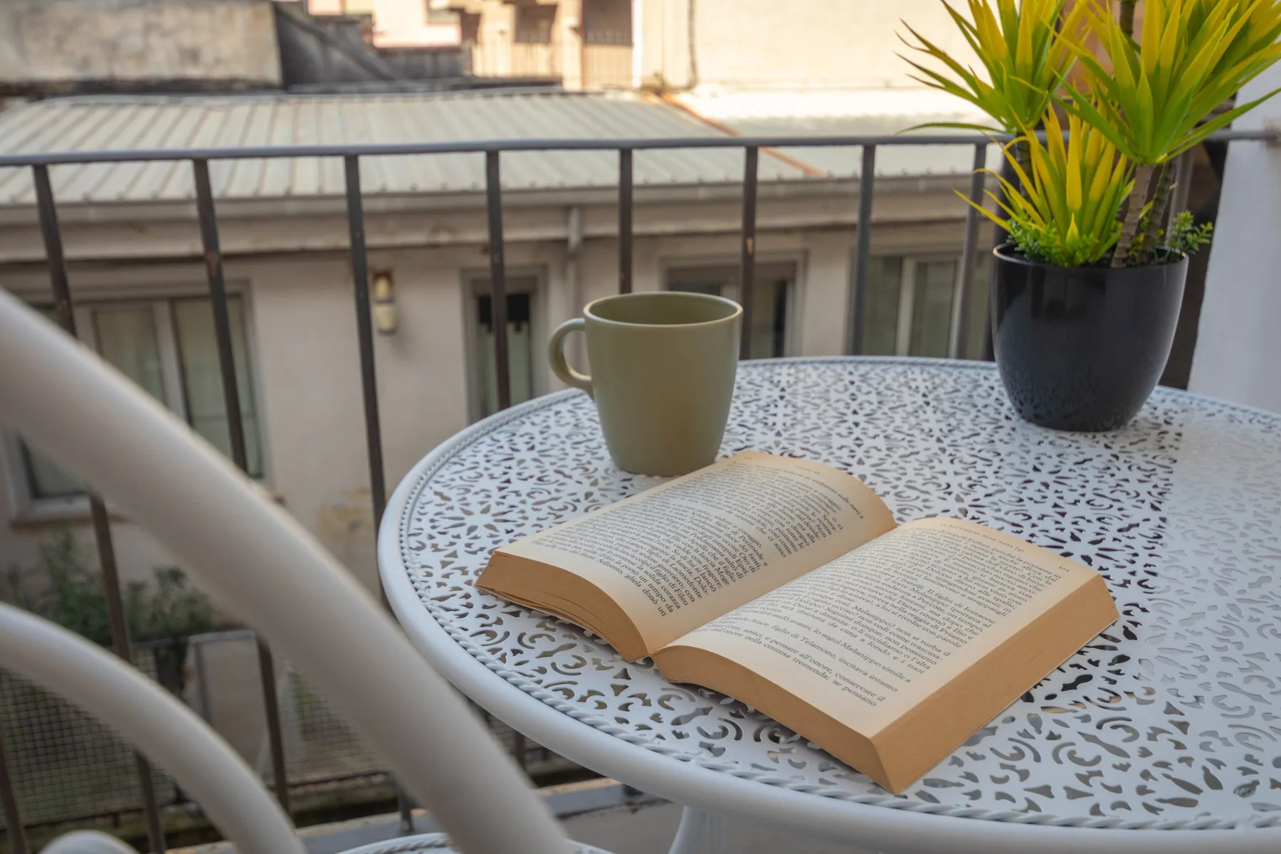 Una mesa de balcón tiene un libro abierto, una taza y una planta en maceta.