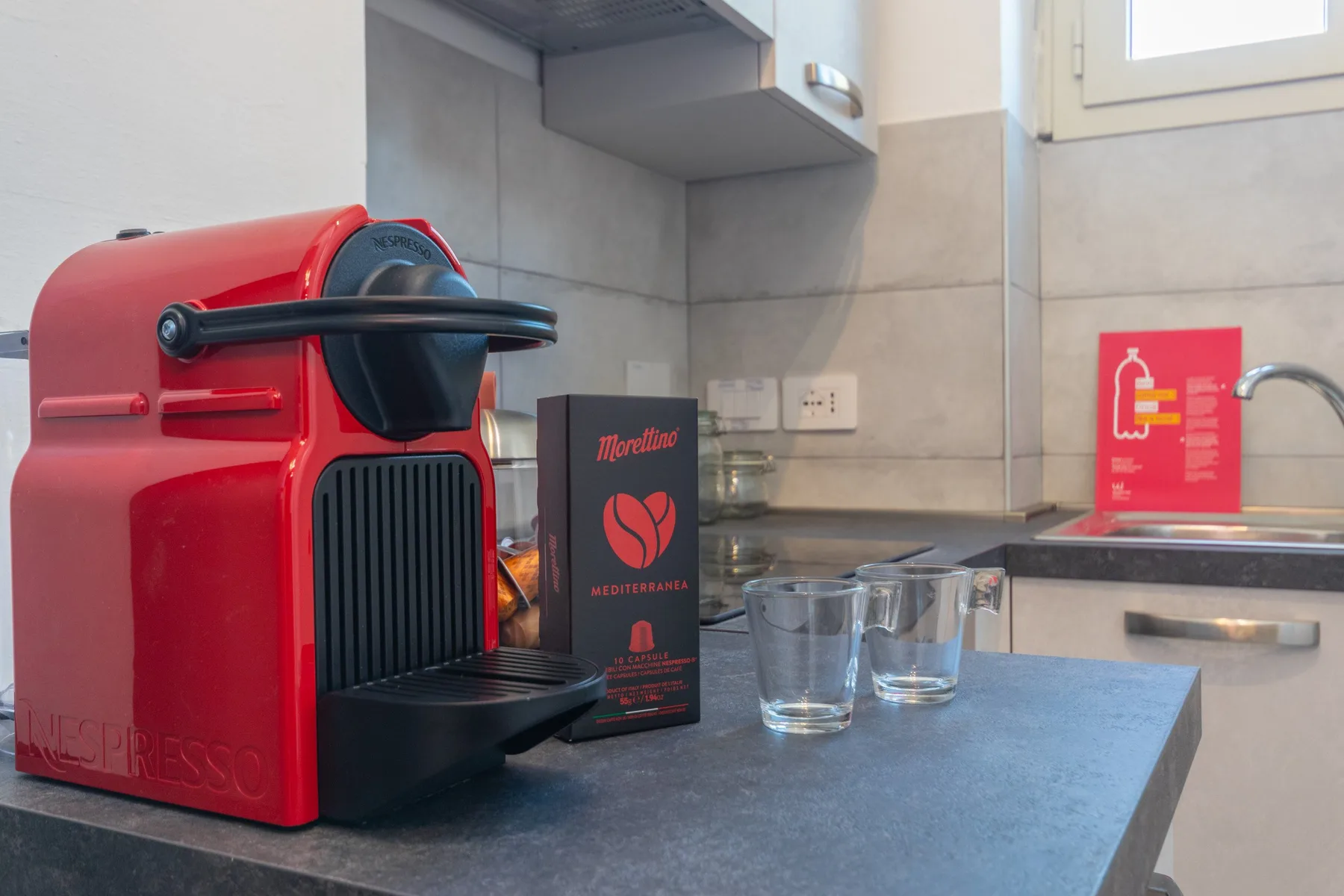 Una encimera de cocina compacta muestra una cafetera roja, cápsulas de café y vasos.