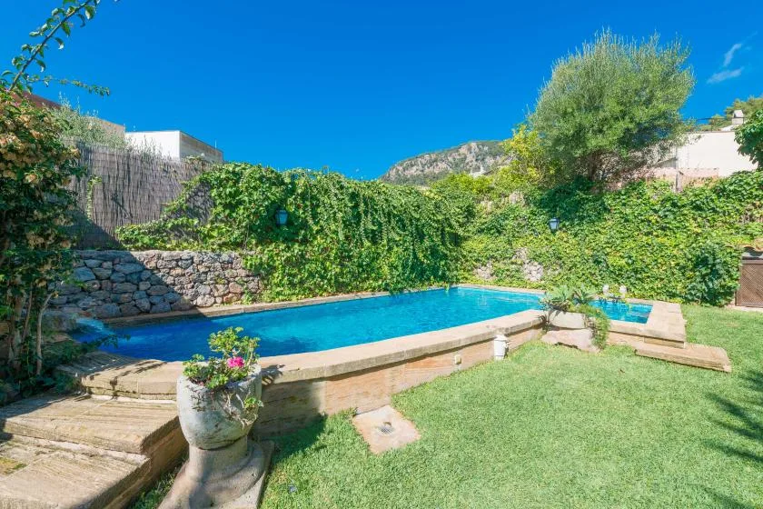 Una piscina al aire libre está junto a un pequeño césped y un jardín frondoso.