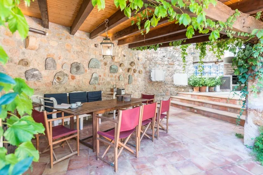 A covered stone patio has a long dining table and leafy plants.