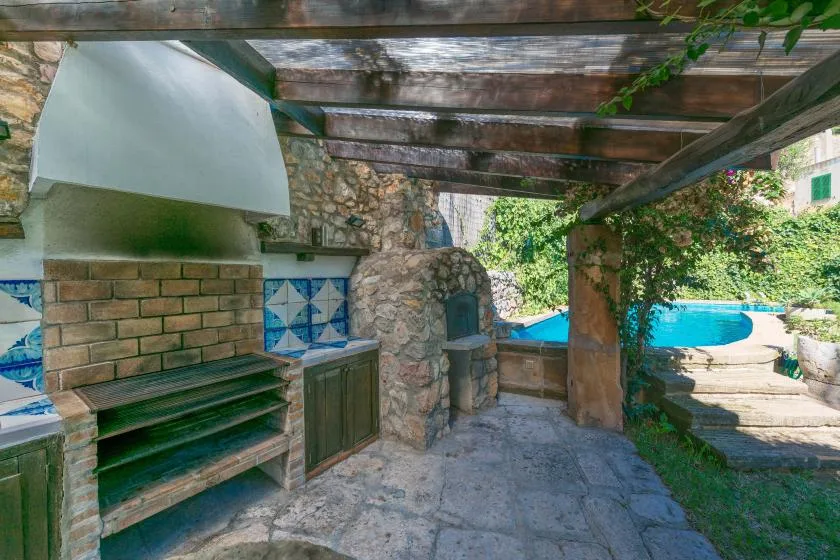 An outdoor patio with a stone barbecue beneath a wooden pergola, beside a pool.