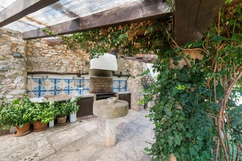 A stone outdoor cooking area with a built-in grill sits beneath a leafy pergola.